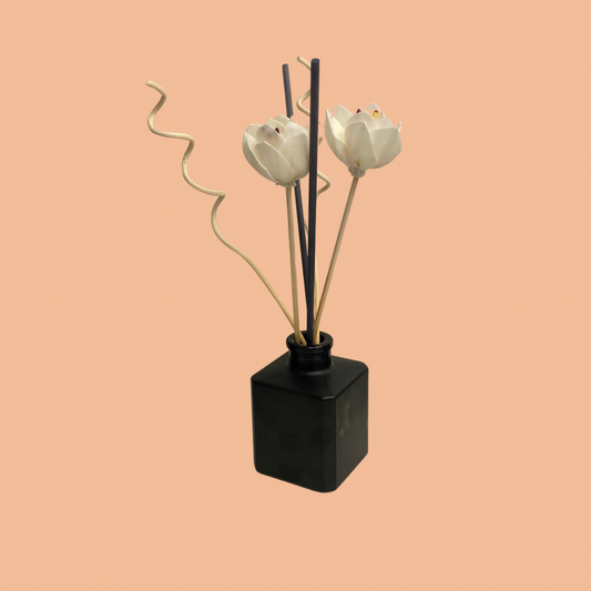stylish reed diffuser paired with a black vase filled with white flowers, set against a soft pink background. The setup exudes elegance and sophistication, adding a touch of charm to your space.