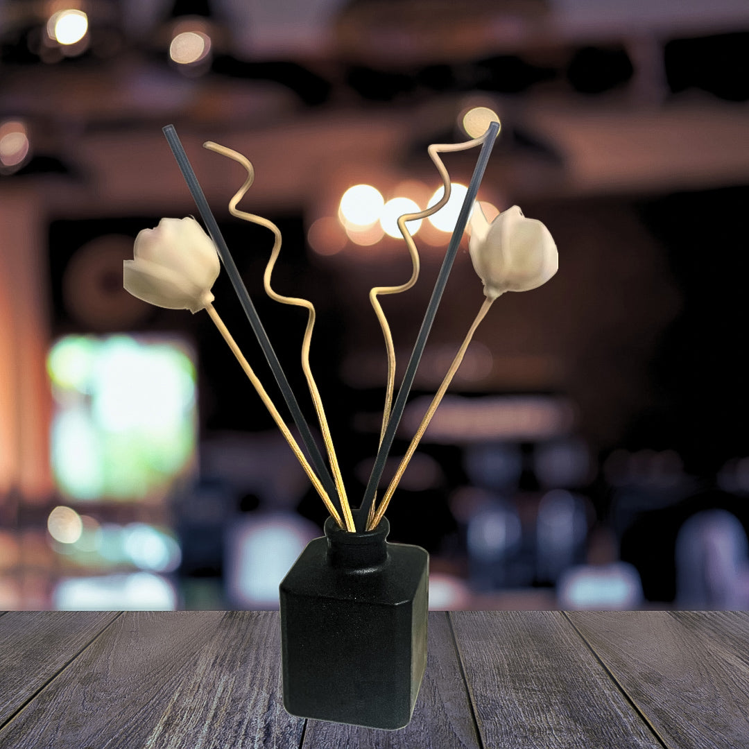 A black vase filled with white flowers and a chic reed diffuser placed on a wooden table, with a blurred cozy home in the background, creating a warm and inviting ambiance.
