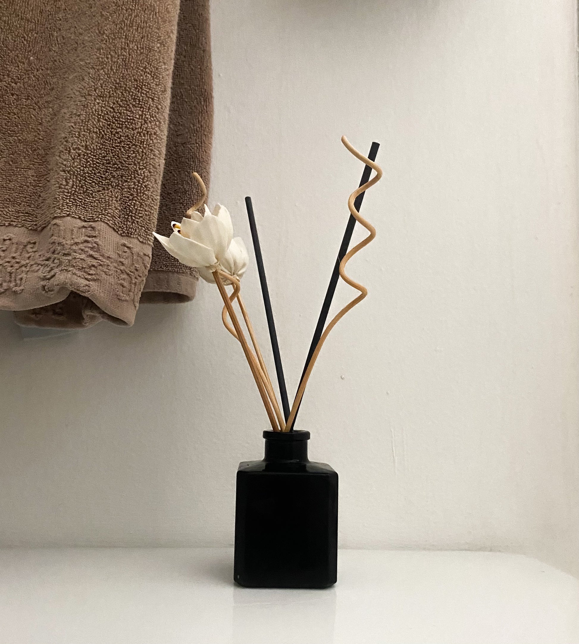 A refined reed diffuser beside a black vase filled with white flowers, placed on a white glass surface with a brown towel hanging behind. The minimalist setup brings a clean and serene vibe to any room.