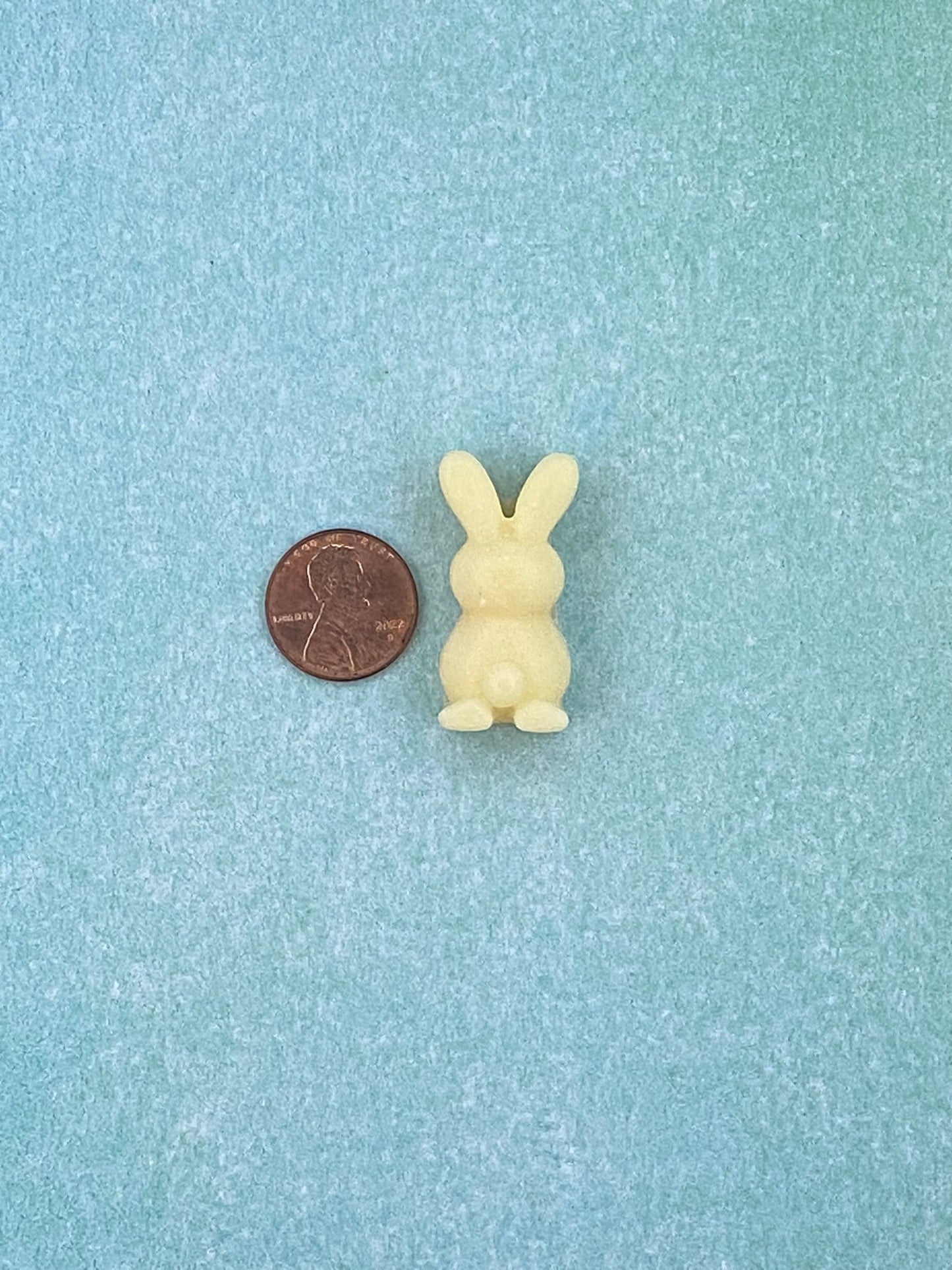 One cream white-colored bunny-shaped soy wax melts beside is a coin. Placed on a soft blue flat surface.