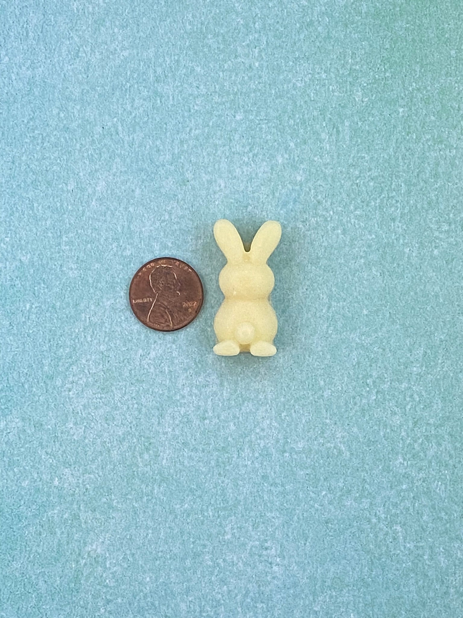 One cream white-colored bunny-shaped soy wax melts beside is a coin. Placed on a soft blue flat surface.