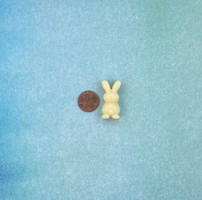 One cream white-colored bunny-shaped soy wax melts beside is a coin. Placed on a soft blue flat surface.