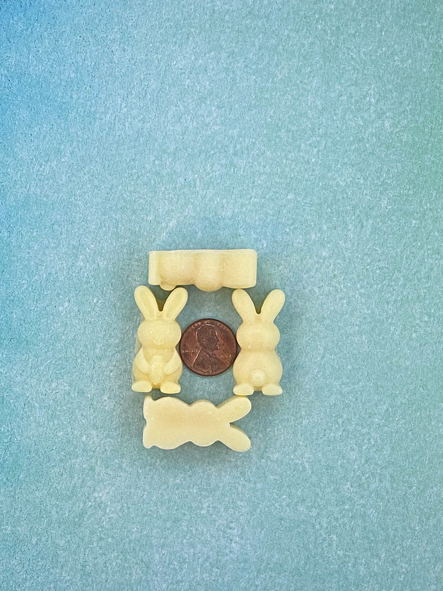 Four cream white-colored bunny-shaped soy wax melts and arranged in four sides with a coin in the middle. Placed on a soft blue flat surface.