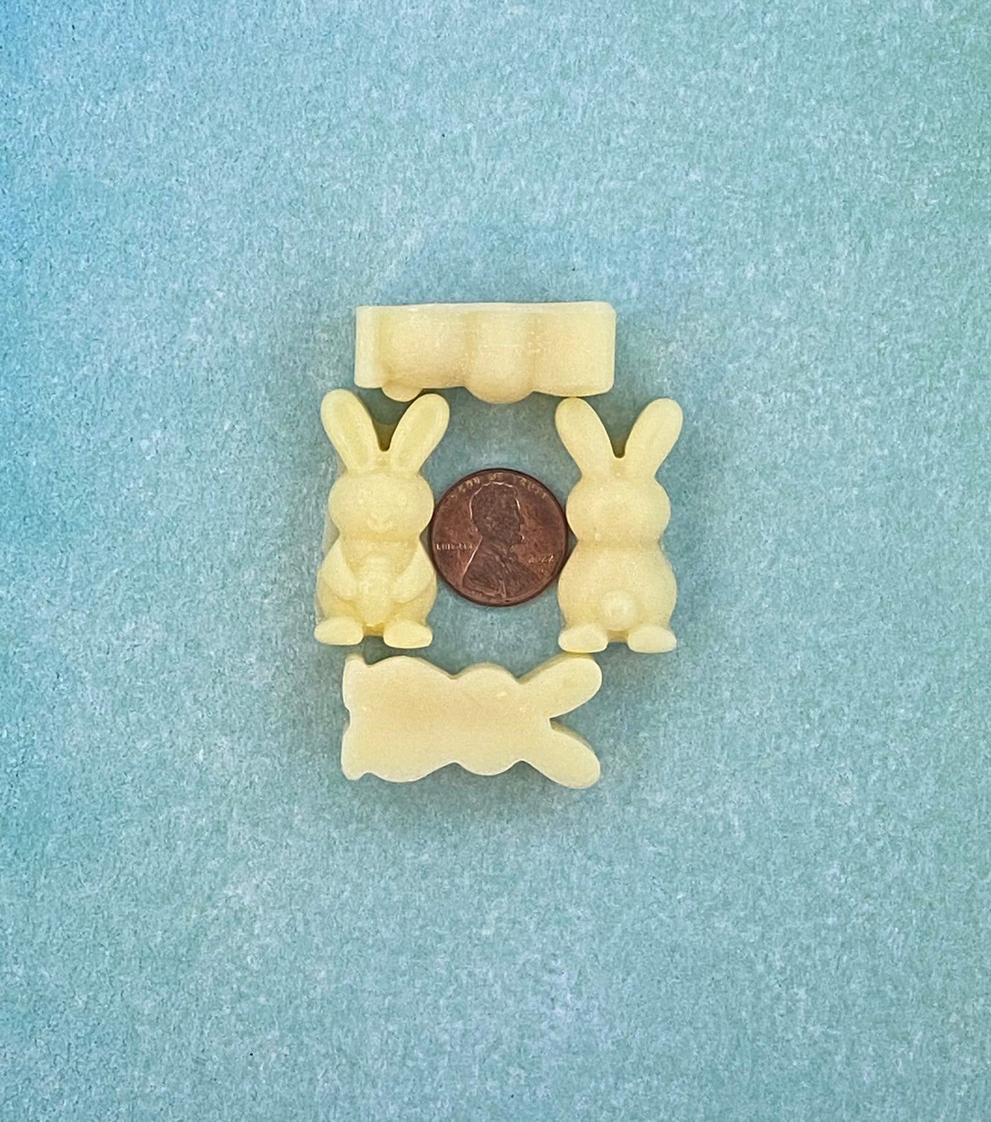 Four cream white-colored bunny-shaped soy wax melts and arranged in four sides with a coin in the middle. Placed on a soft blue flat surface.