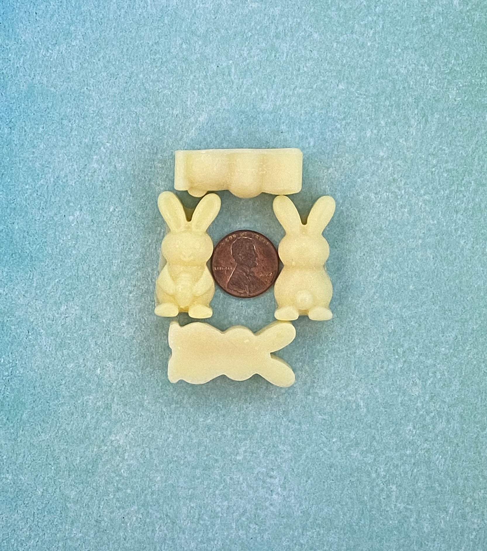 Four cream white-colored bunny-shaped soy wax melts and arranged in four sides with a coin in the middle. Placed on a soft blue flat surface.