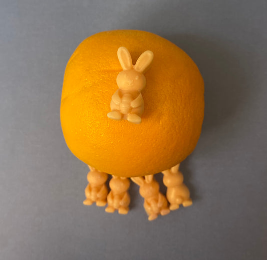 An orange bunny-shaped soy wax melt candle perched on top of a fresh orange fruit, with four additional orange bunny-shaped candles scattered below on a grayish flat surface. The setup blends fruity freshness with a whimsical touch.