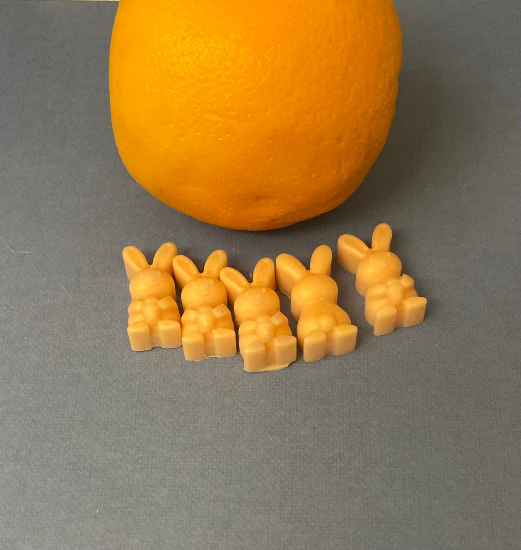 Five orange bunny-shaped scented soy wax melts arranged on a light purple flat surface, with a large orange apple placed behind them, creating a vibrant and playful contrast of colors.