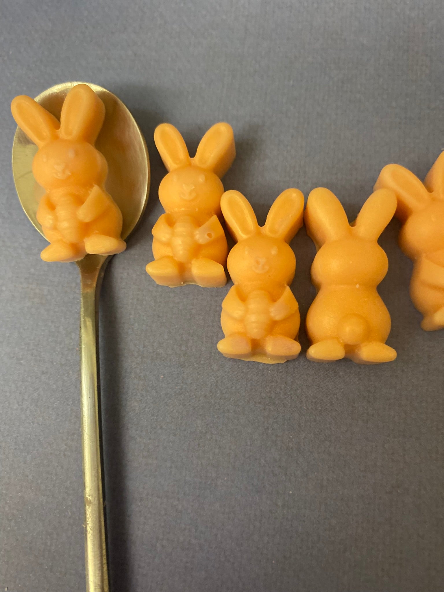 Five orange bunny-shaped scented soy wax melts arranged on a light purple flat surface, one is scooped on a golden spoon.