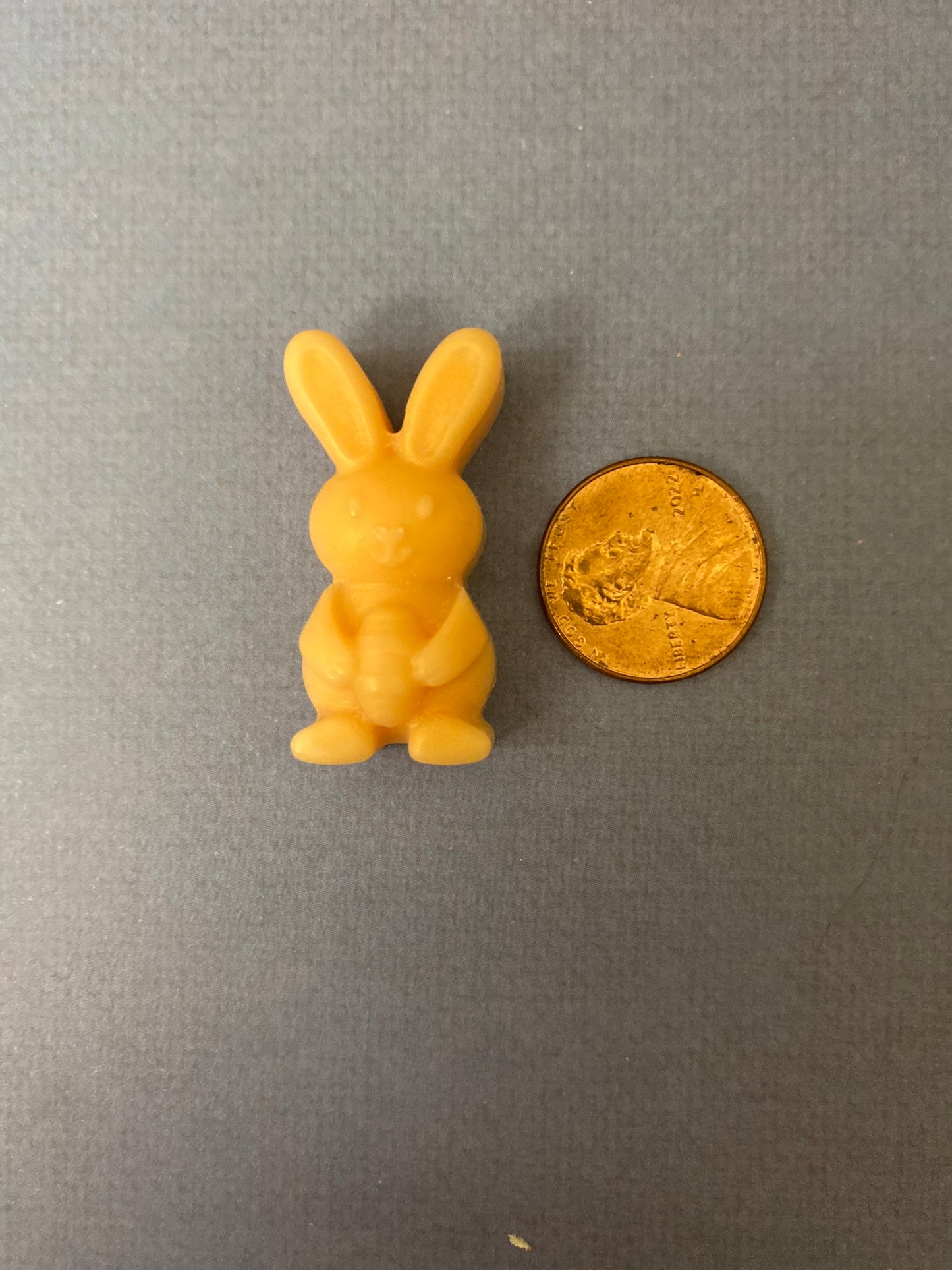 One orange bunny-shaped scented soy wax melts sitting beside a golden coin on a light purple flat surface.
