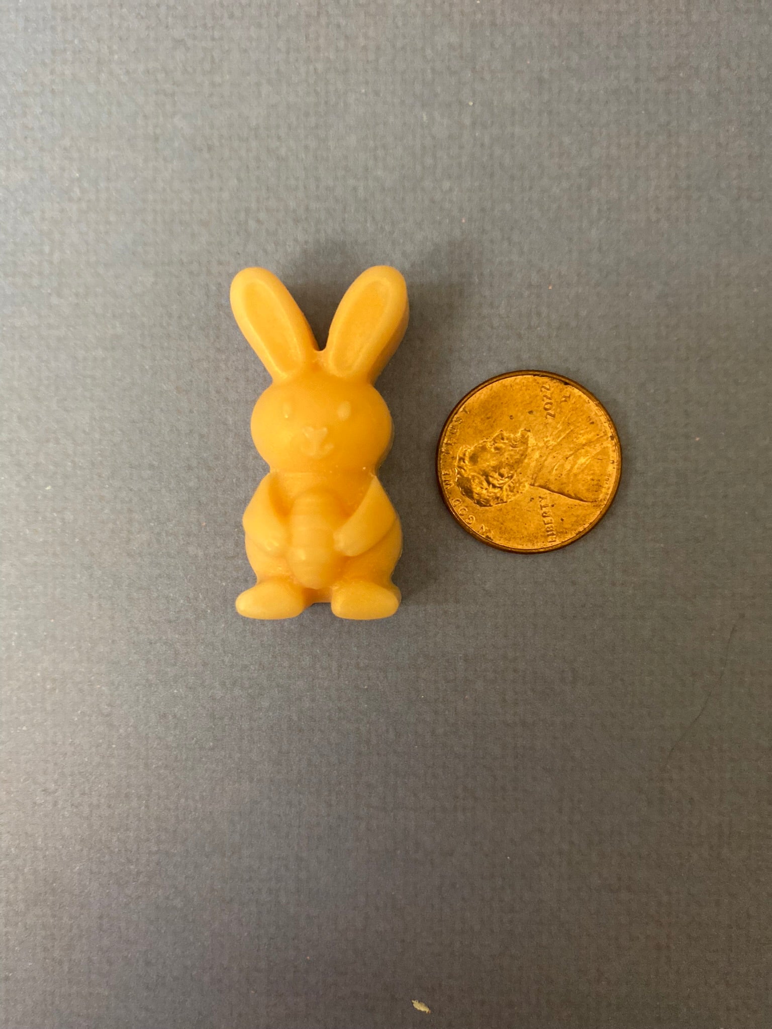 One orange bunny-shaped scented soy wax melts sitting beside a golden coin on a light purple flat surface.