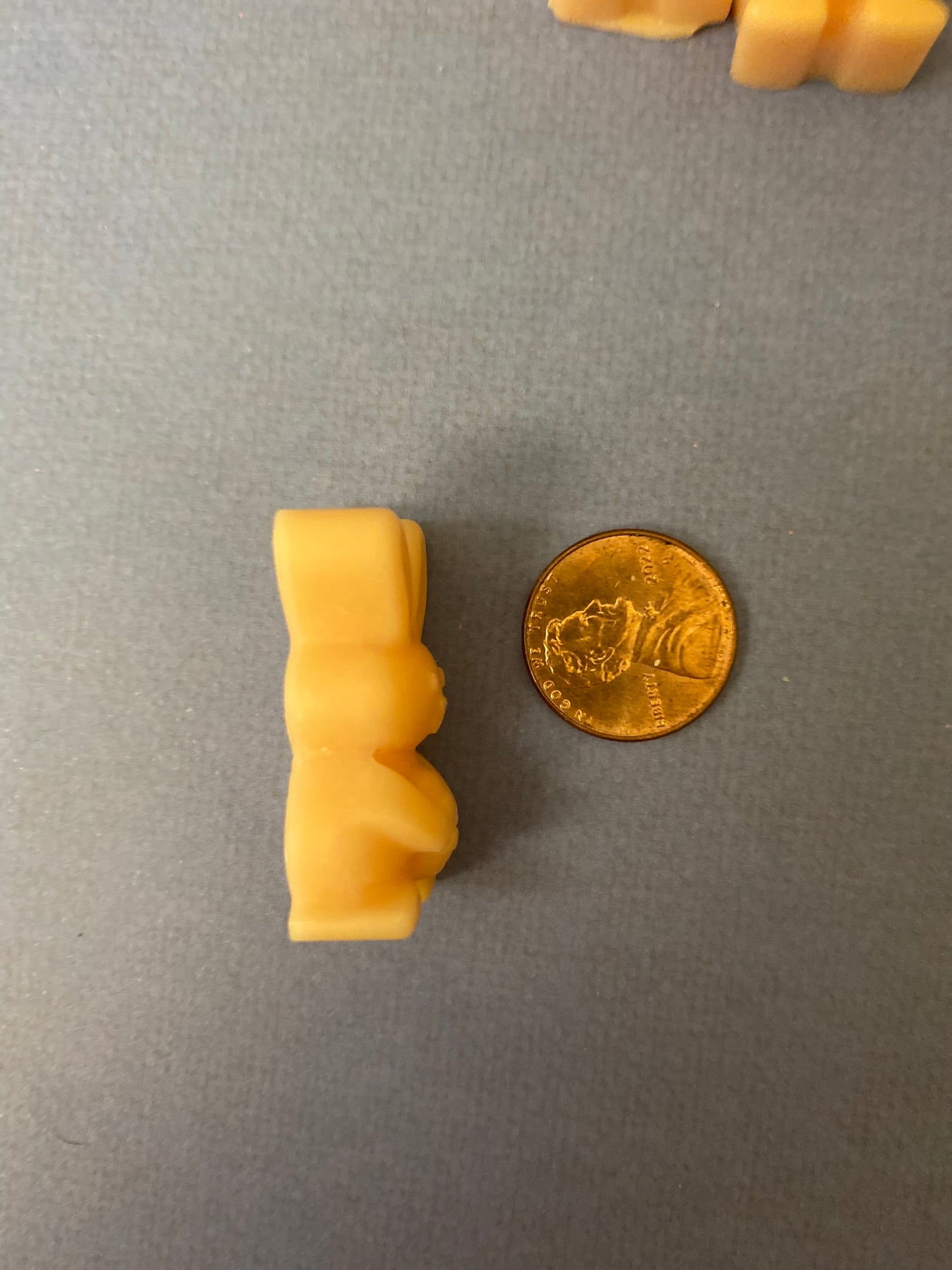 One orange bunny-shaped scented soy wax melts laying on its side facing a golden coin on a light purple flat surface.