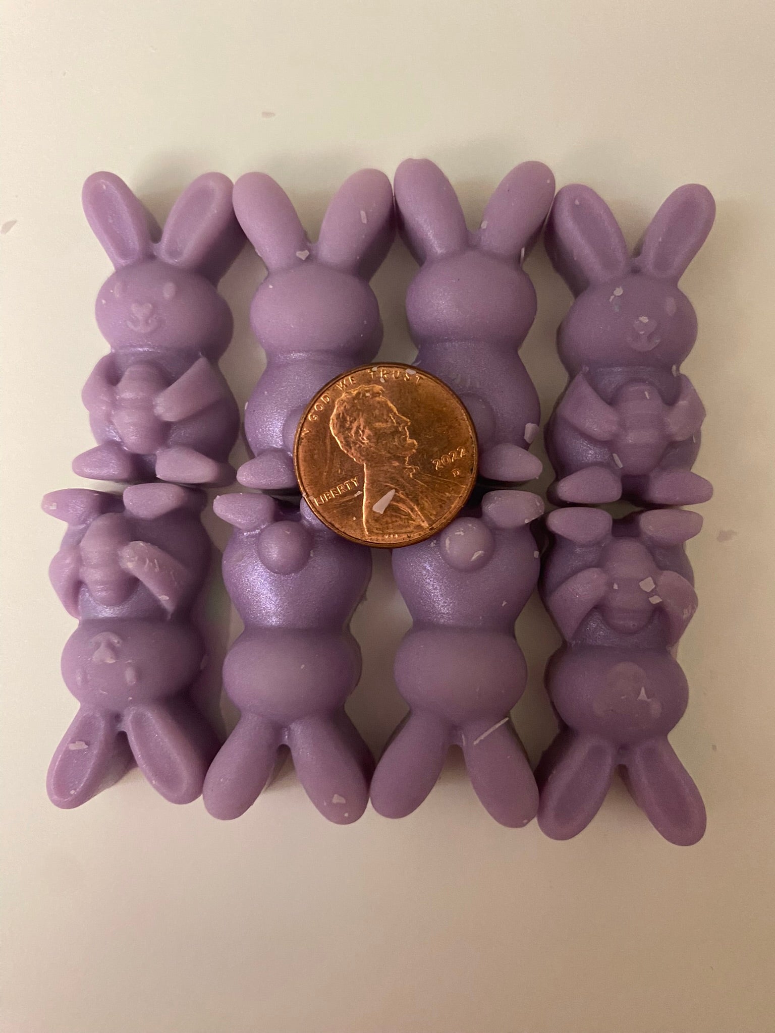 Eight purple-colored bunny-shaped soy wax melts with a golden coin on top arranged on a light pink flat surface, creating a vibrant and cheerful display that embodies playful and uplifting vibes.