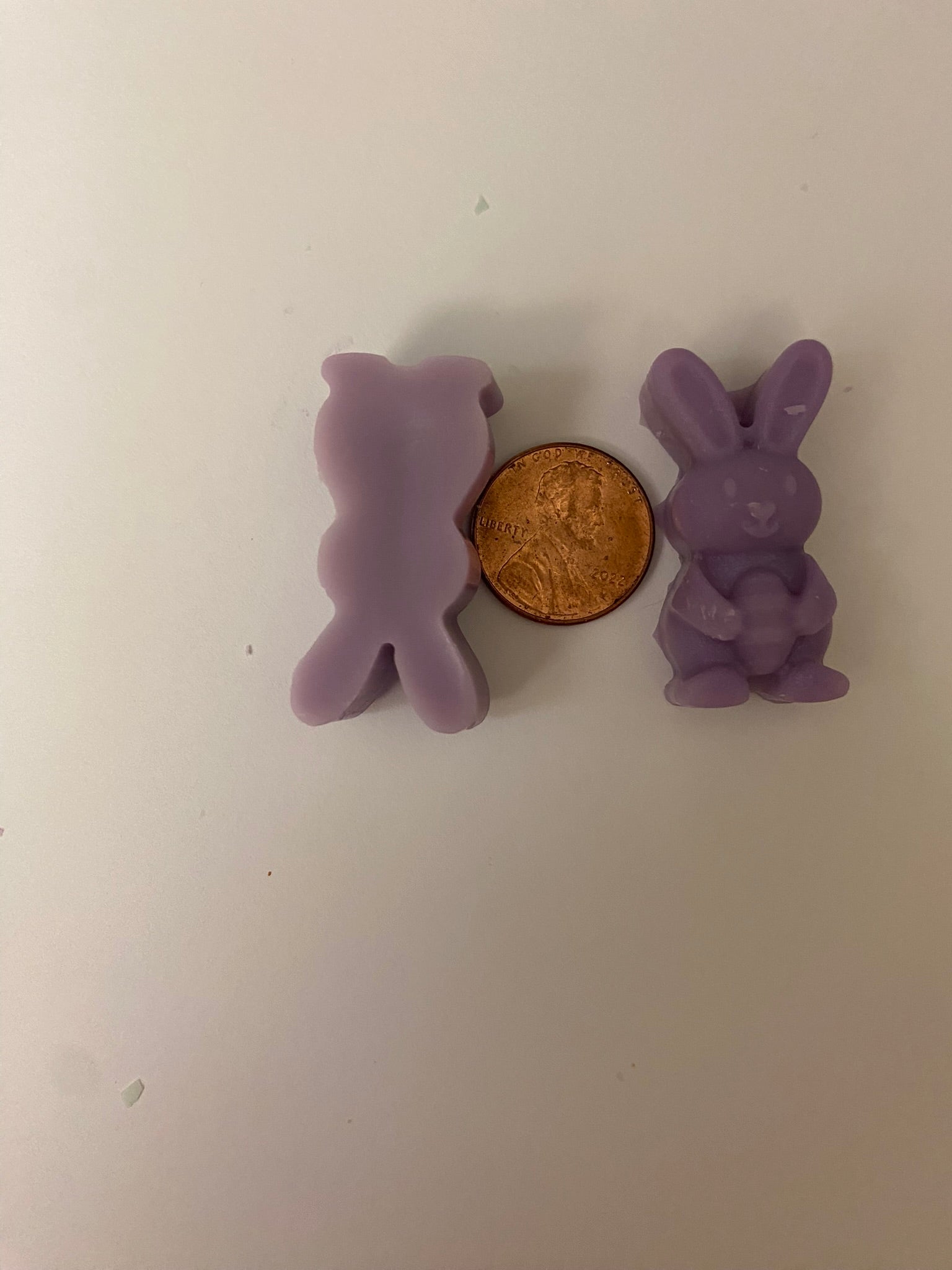 Two purple bunny-shaped soy wax melts with a golden coin in between. One bunny is facing upwards and the other downwards, arranged on a light pink flat surface.