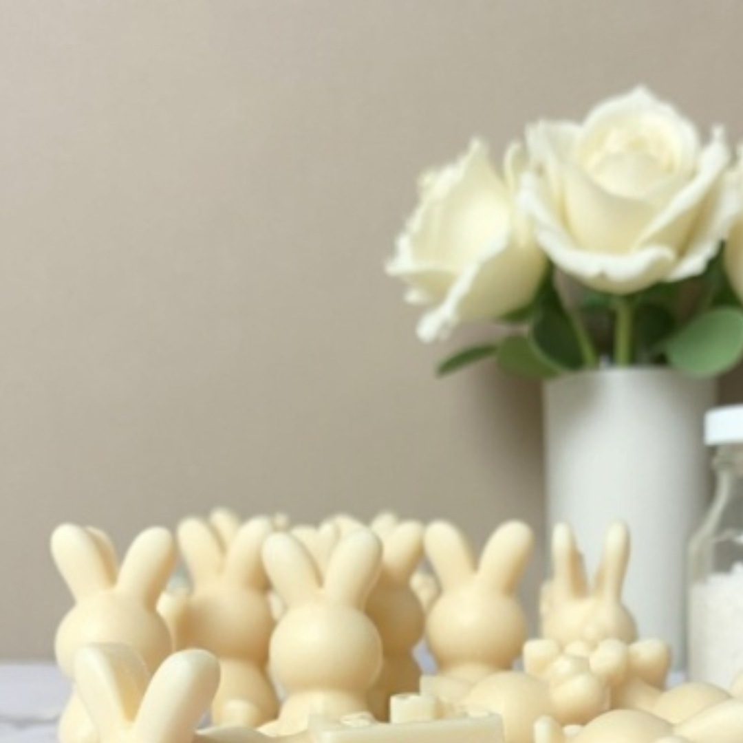 A set of creamy vanilla-colored bunny-shaped wax melt scented candles, standing in front of a vase of white roses.