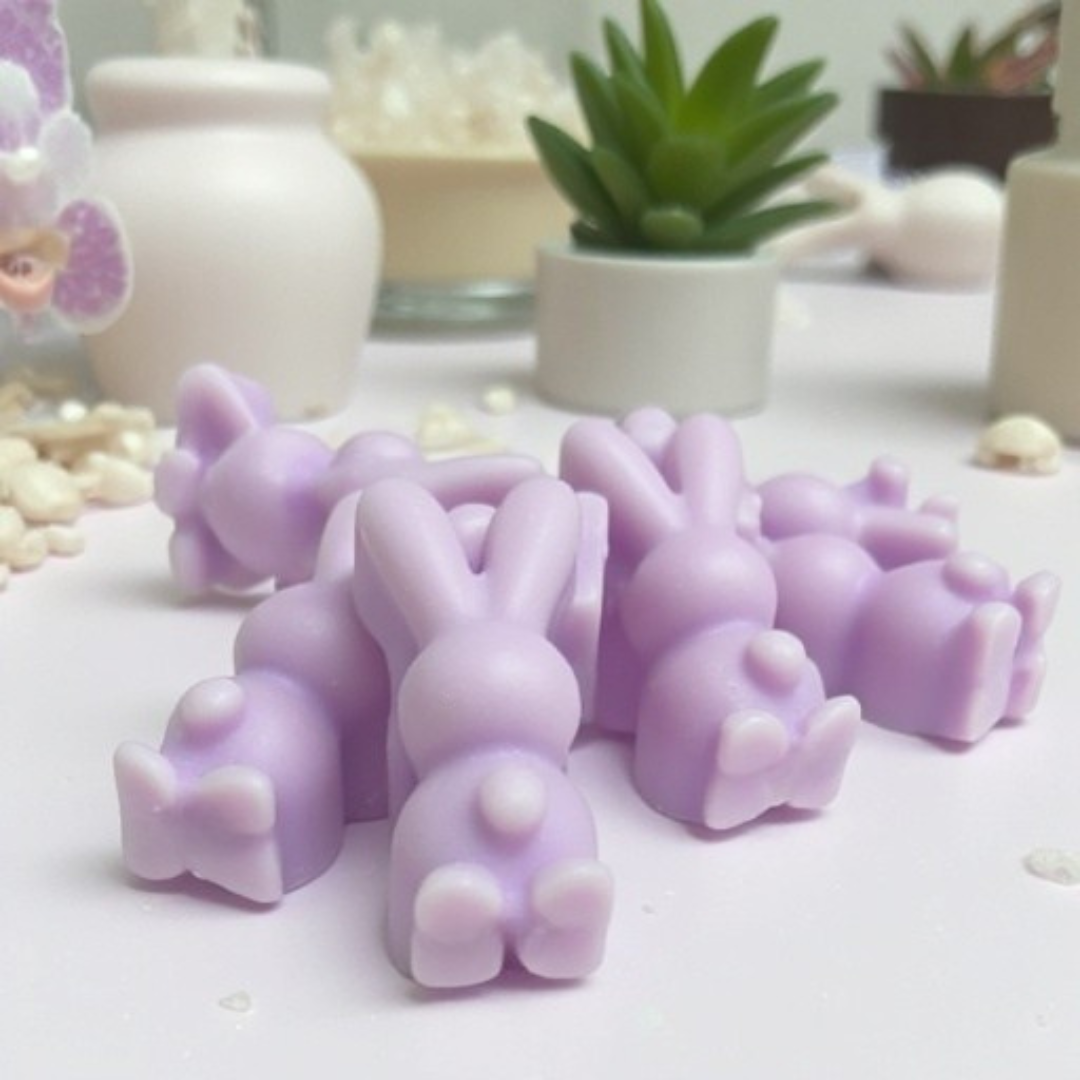 Bulk pieces of light purple bunny-shaped soy wax melts with an orchid flower in the background. A set of mini vases with small plants are blurred in the background.