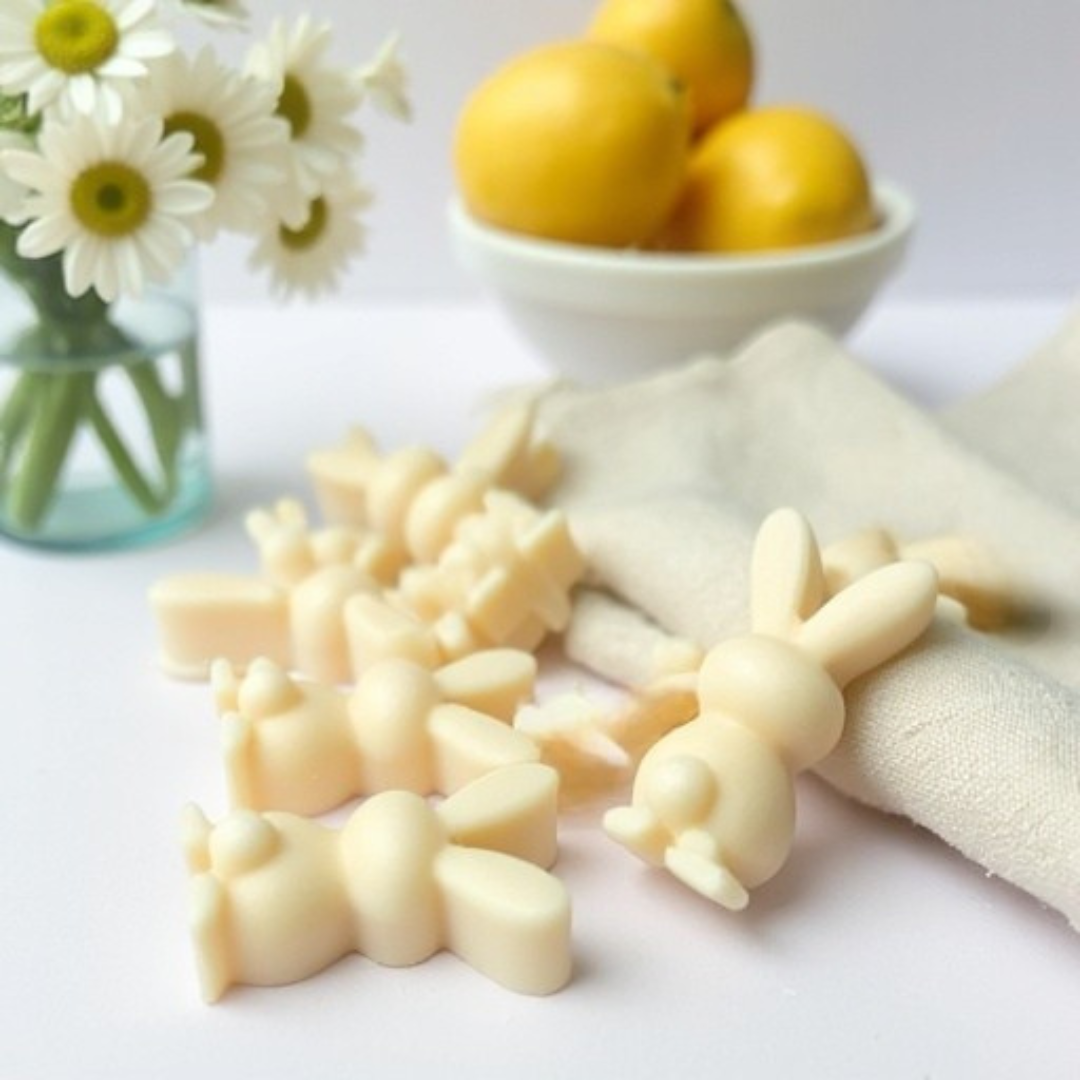 Multiple bunny-shaped Sun-Kissed Linen wax melts arranged on a cream-pink flat surface, with a bowl of lemon fruit and a vase of white flowers in the background, leaning on folded white towels.