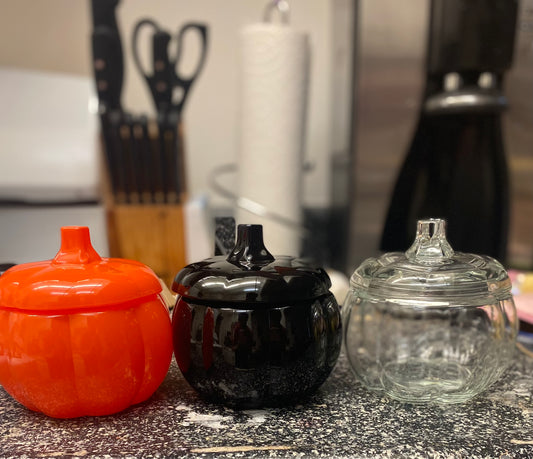 three glass, pumpkin shaped candle jars sit in a row on a kitchen counter. One orange, one black, and one clear. 