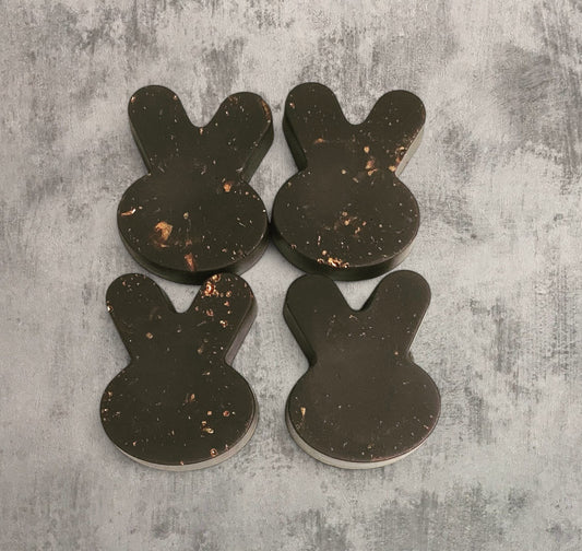 Four black currant-colored bunny-shaped wax melts with marbled gold details on top of a flat grey surface.