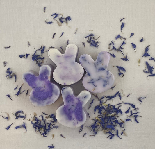 Four lavender-colored bunny-shaped wax melts with marbled purple details, surrounded by scattered dried purple flower petals on a neutral background.