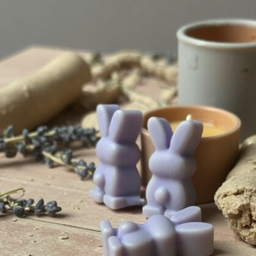 Close-up of lavender bunny-shaped wax melts alongside a lit candle and a piece of driftwood, with dried lavender sprigs and natural decor pieces creating a cozy, calming atmosphere.