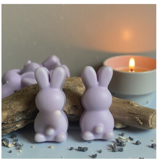 two purple, bunny shaped wax melts leaning against 2 pieces of driftwood with a small candle burning in the background