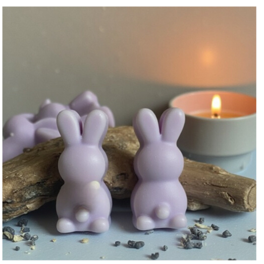 two purple, bunny shaped wax melts leaning against 2 pieces of driftwood with a small candle burning in the background