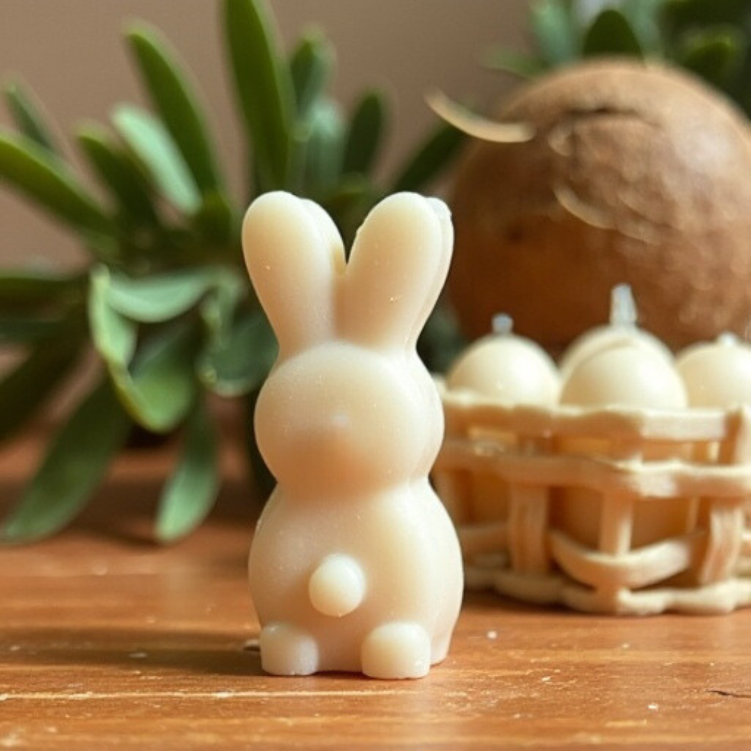 One cream colored, small bunny shaped wax melt standing on a clean wood table top with a plant and coconut in the background