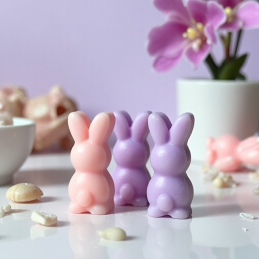 Three bunny-shaped soy wax melts in pink, dark purple, and light purple, placed beside a white bowl with sea salt scattered around them. An orchid flower in a vase is positioned in the background.